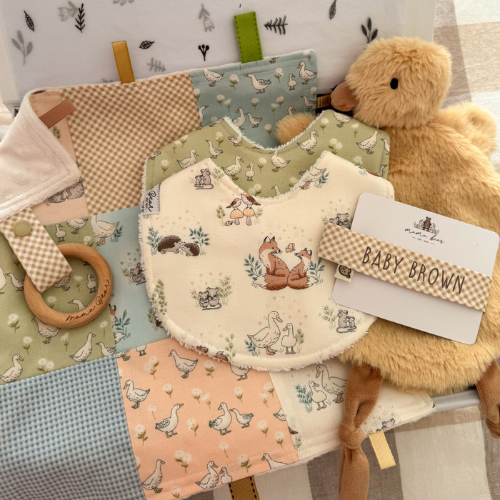 Personalised baby gift box featuring handmade cotton baby bibs in woodland and duck prints, a personalised gingham dummy clip, yellow duck comforter, wooden teething ring with Taggie comforter and coordinating baby accessories in soft shades of blue, peach, green and neutral tones.