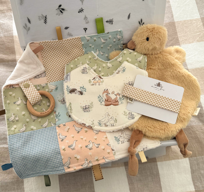 Personalised unisex baby gift box featuring a handmade Taggie Blankie with woodland fox, duck, bunny and gingham fabric squares, personalised gingham dummy clip, coordinating handmade baby bibs in woodland and duck prints, and a soft plush duck comforter in pale yellow. Fabrics are in shades of blue, peach, green and neutral tones.