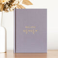 Photo of a pregnancy journal in a dark grey linen shade. The front of the book as gold writing on the front that says 'Hello Little Mama'.