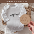 White baby onesie with 'surprise!' text and a wooden disc reading 'We're having a baby' on a textured surface.