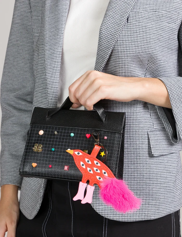Close-up of a black top-handle handbag with a grid pattern, colorful studs, and a playful red bird charm with multiple eyes and a pink tail, held by a person in a gray blazer.