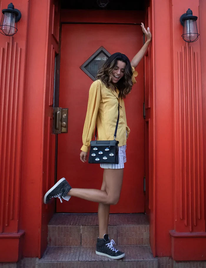 Woman in a yellow blouse and striped shorts poses in a doorway with a bright red frame, wearing a black crossbody bag with eye motifs and lifting one leg playfully.
