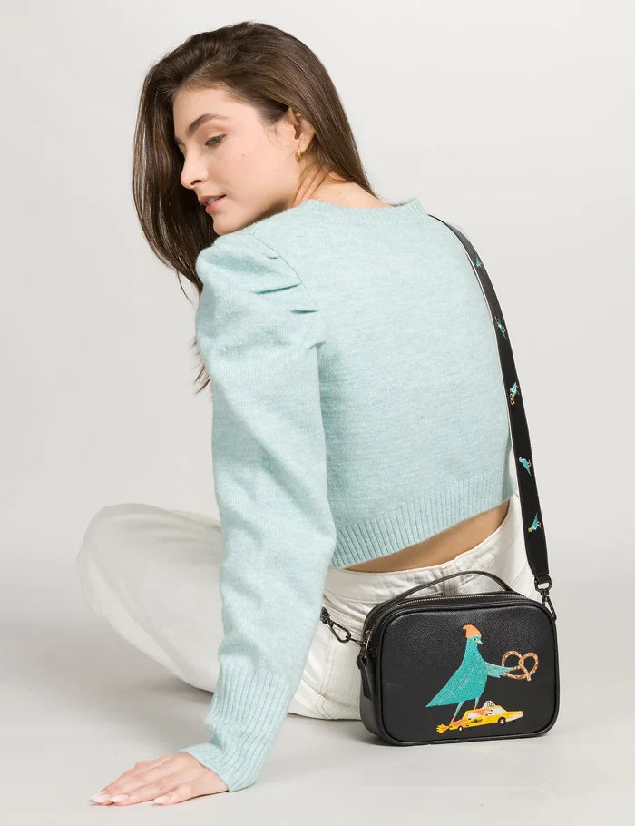 Woman in a light blue cropped sweater and white pants sits turned away, looking over her shoulder while wearing a black crossbody bag with a colorful embroidered bird design.