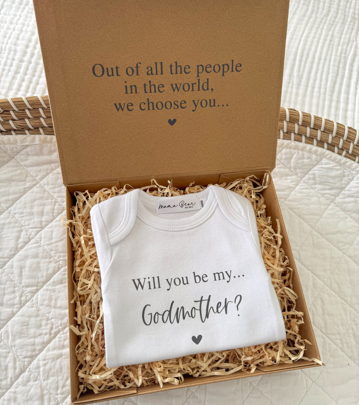 Kraft gift box with black text on inside lid "Out of all the people in the world, we choose you..." inside box is raffia and a white baby onesie saying ' WIll you be my... Godmother?" Onesie can be customised to also say Godfather or Godparents